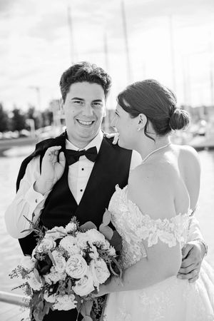 Black and white photo of smiling groom in tuxedo and bride in lace wedding dress holding bouquet near marina in Hamilton