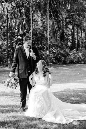 Black and white photo of a bride sitting on a swing holding a bouquet while the groom stands beside her outdoors.