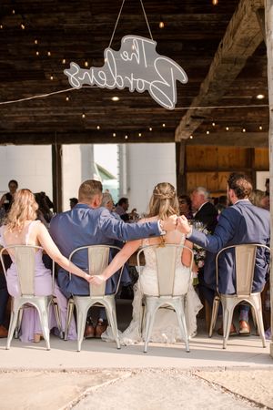 Bride and groom seated at reception table under neon sign with wedding party holding hands, at Brantview in St. George.