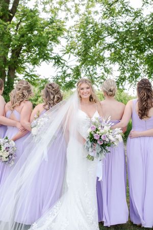 Smiling bride in white gown holding bouquet surrounded by bridesmaids in lavender dresses outdoors, at Brantview in St. George.