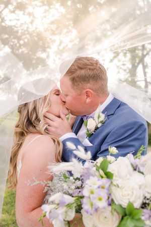 Bride and groom kissing under a flowing veil with a white and purple bouquet in the foreground, at Brantview in St. George.