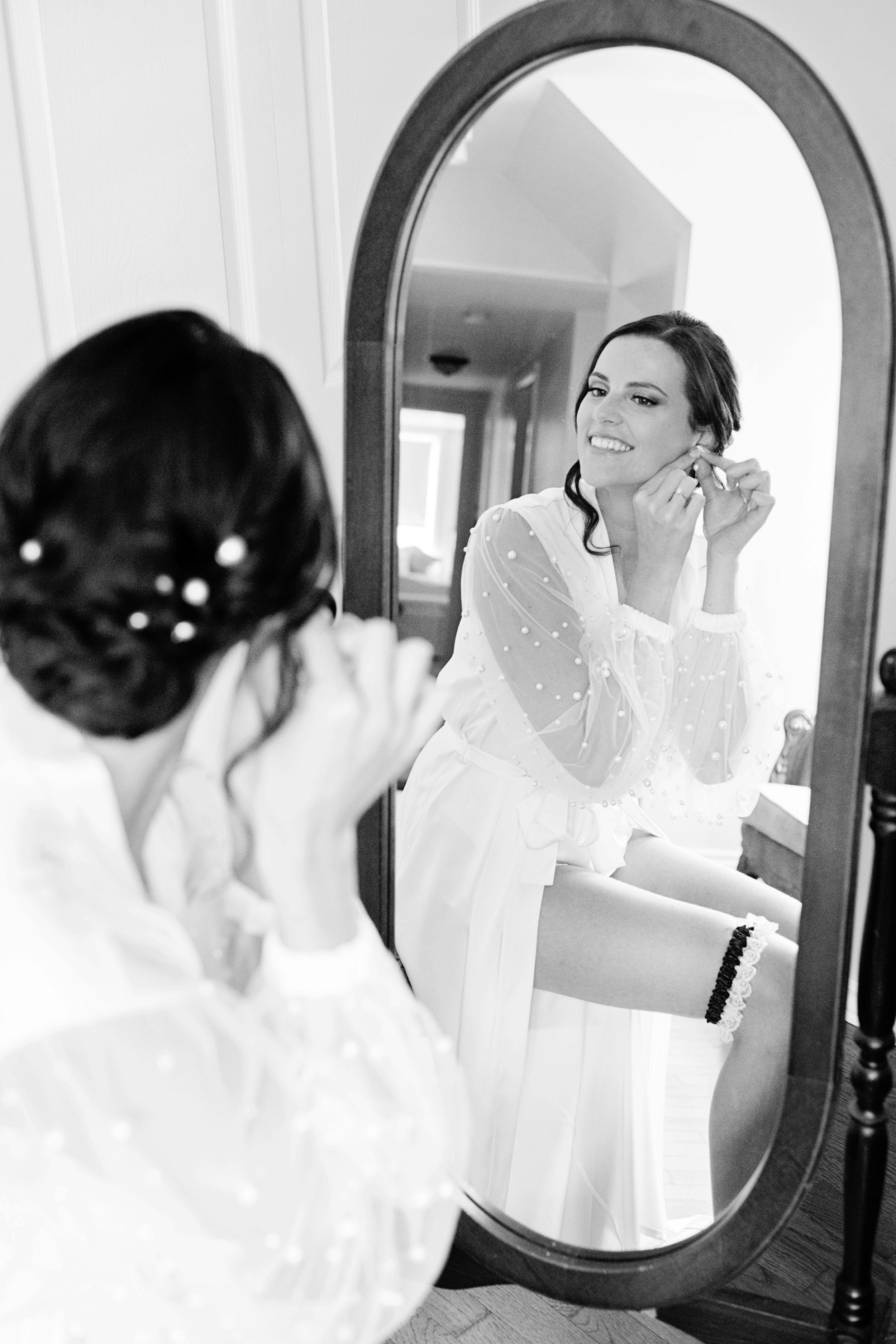 Bride putting on earrings while getting ready, reflected in an elegant oval mirror in black and white.