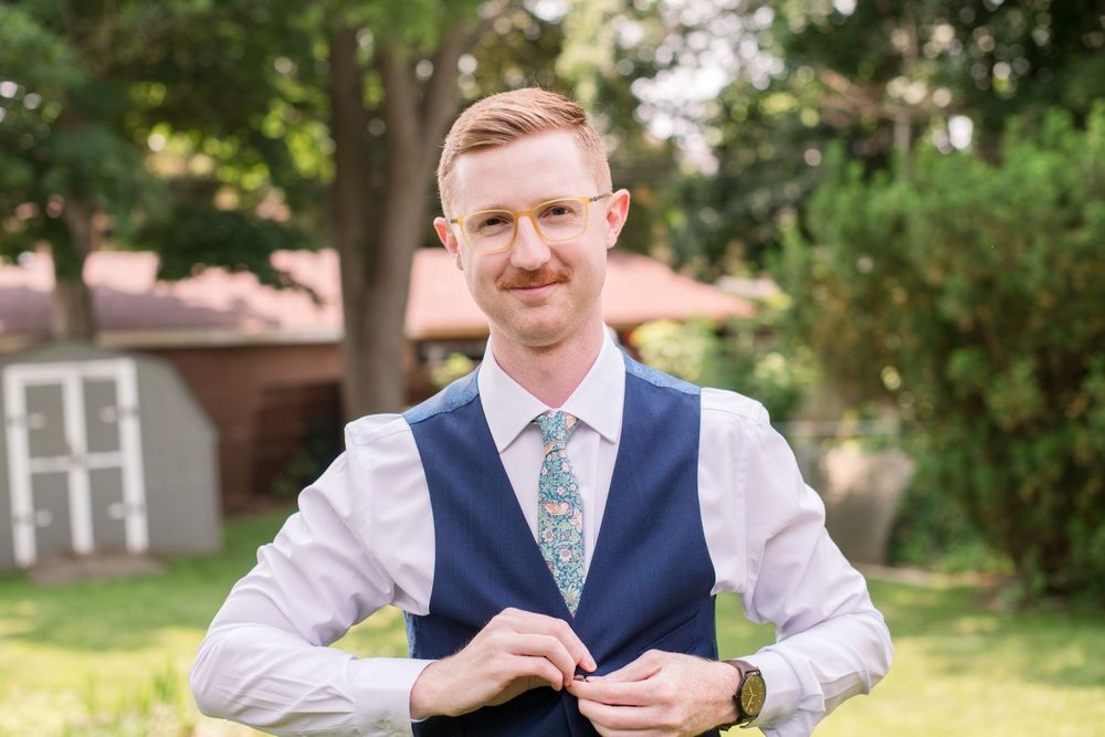 Smiling groom in navy vest and floral tie buttons his jacket while standing outdoors before the wedding , In Paris Ontario.
