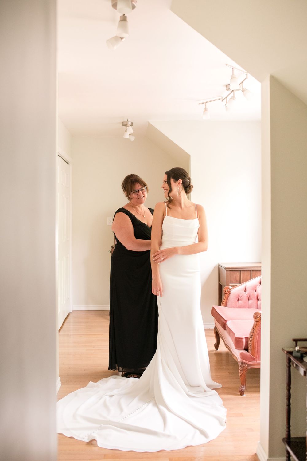 Mother helps bride in sleek white gown prepare in a bright, elegantly decorated dressing room.