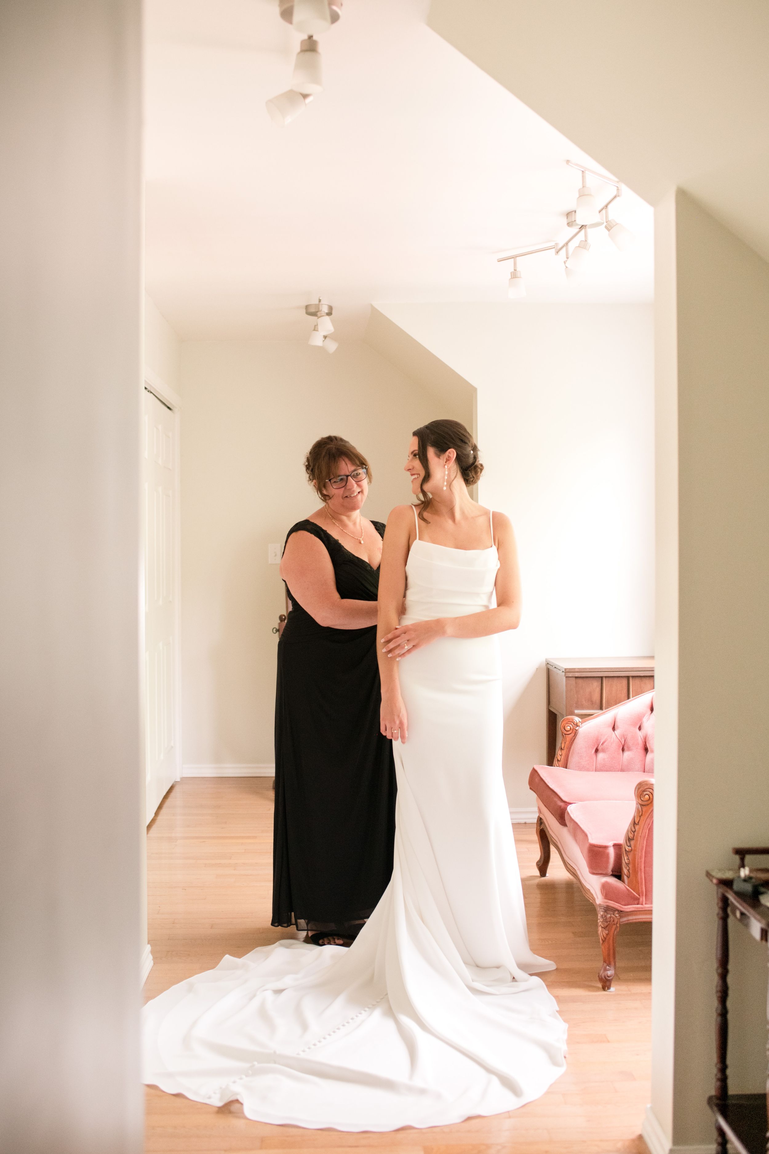 Mother helps bride in sleek white gown prepare in a bright, elegantly decorated dressing room.