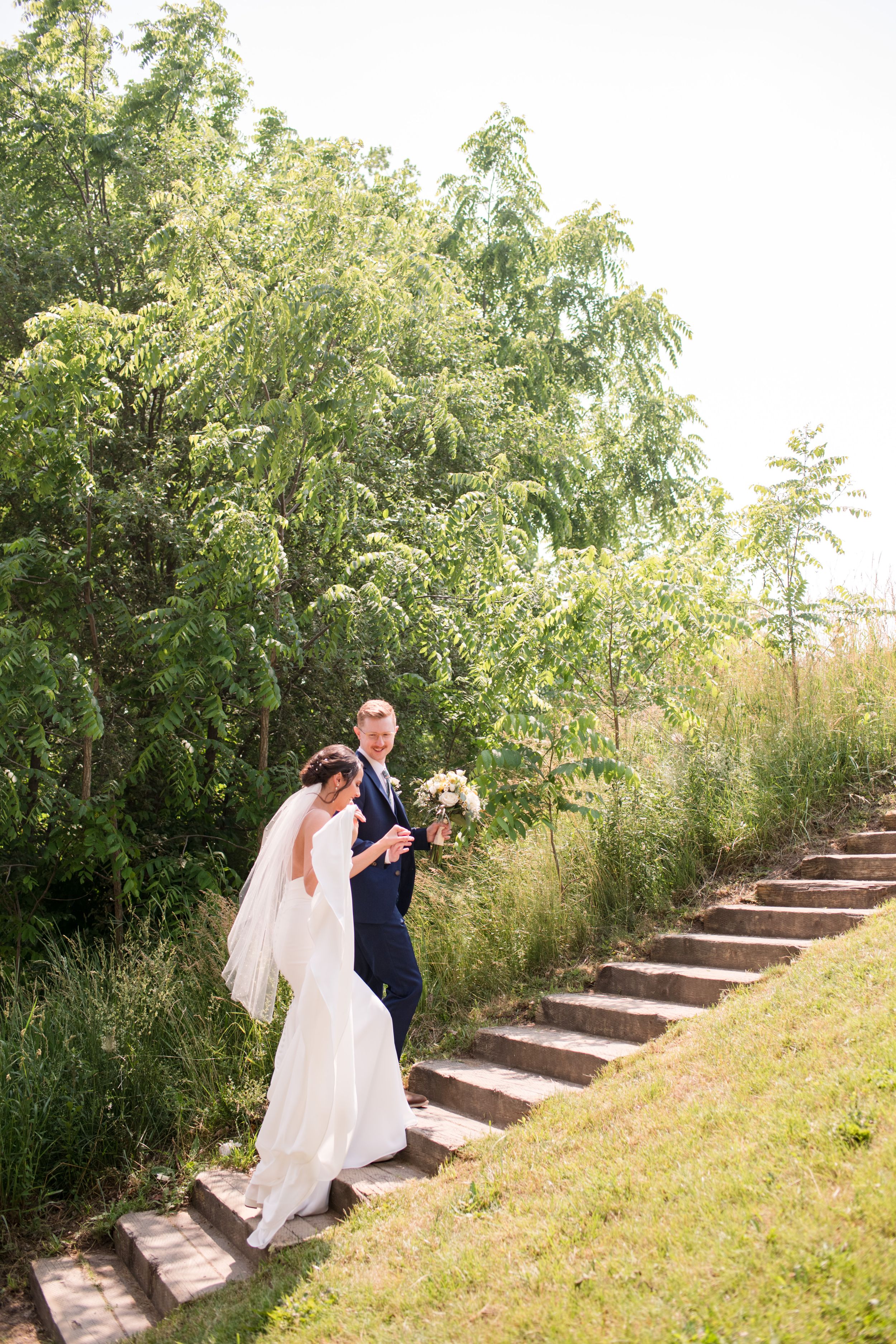 Bride and groom walk hand in hand up stone steps surrounded by lush green trees and tall grass., Paris Ontario.