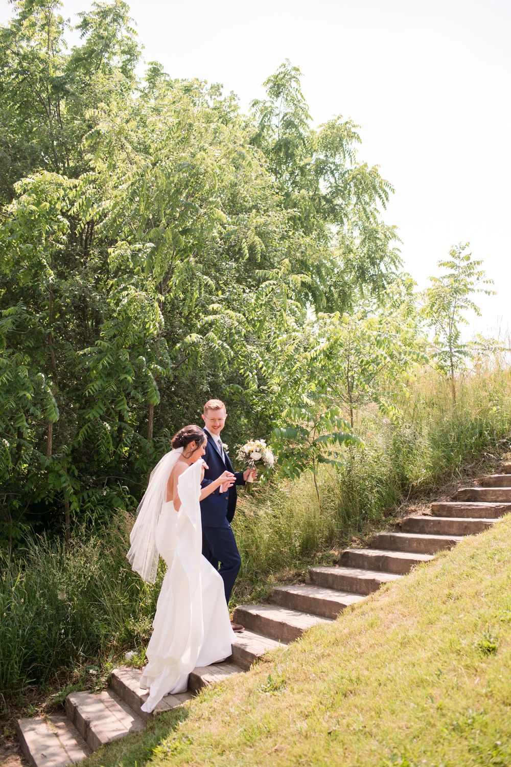 Bride and groom walk hand in hand up stone steps surrounded by lush green trees and tall grass., Paris Ontario.
