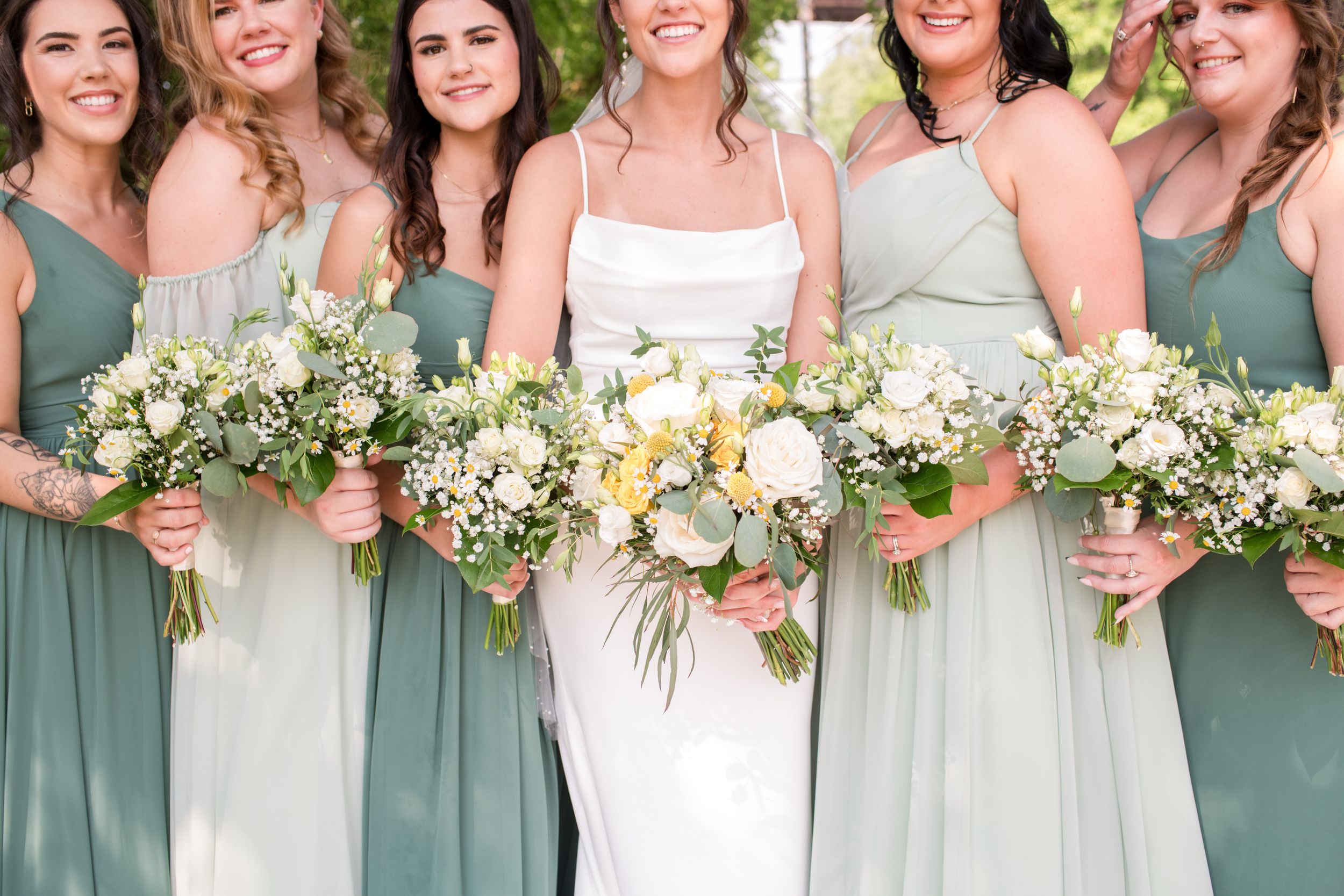 Bride in white gown poses with bridesmaids in sage green dresses holding elegant floral bouquets.
