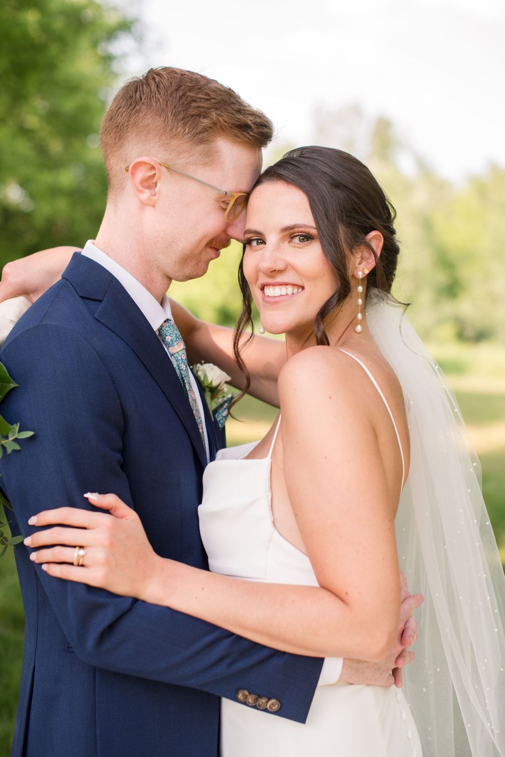 Smiling bride in white gown embraces groom in navy suit during outdoor wedding portraits in a garden.