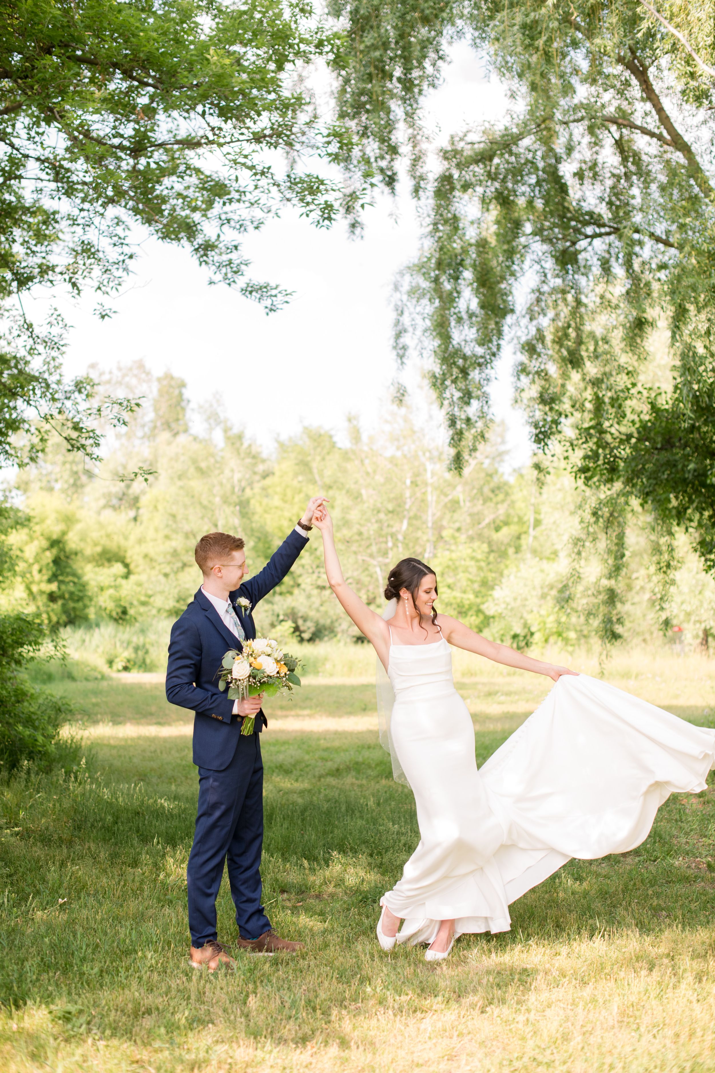 Joyful groom spins bride in flowing white dress under a willow tree in a lush green outdoor setting.
