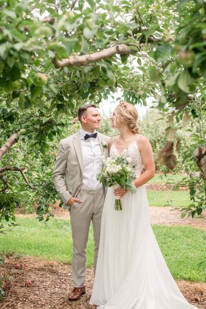 Bride and groom gazing at each other under lush apple tree branches, bride holding white bouquet.
