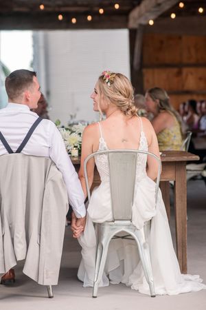 Bride and groom hold hands at sweetheart table in rustic barn reception with string lights overhead at Brantview in St. George.
