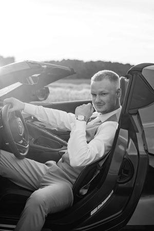 Dapper groom in tan vest and white shirt sits in a luxury convertible car in a black and white portrait.