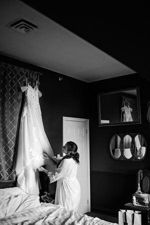 Bride in white robe reaches for lace wedding gown hanging in dark elegant hotel room in Paris Ontario.