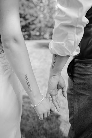 Close-up black and white photo of bride and groom holding hands revealing matching Roman numeral date tattoos on their forearms.