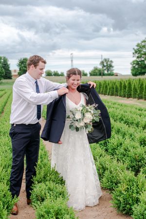 Groom places his navy blazer over smiling brides shoulders in lush green garden rows.