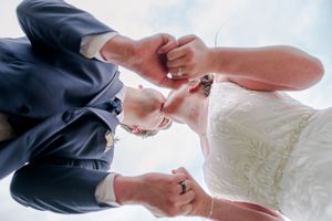 Low angle view of bride and groom holding hands with wedding rings visible against blue sky.