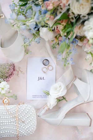 Elegant flat lay of wedding details including white heels, bouquet, pearl clutch, gold rings, and J&L monogram card.