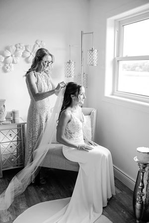 Black and white photo of mother carefully placing wedding veil on seated bride near a bright window while getting ready.