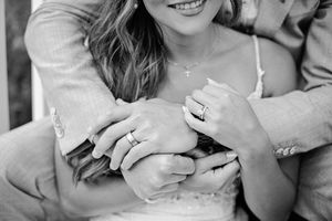 Close-up black and white photo of newlyweds hands intertwined showing wedding rings and a delicate cross necklace.