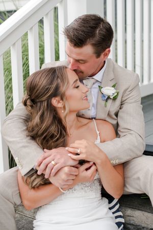 Groom tenderly embraces bride from behind as they sit together on porch steps in an intimate wedding portrait.