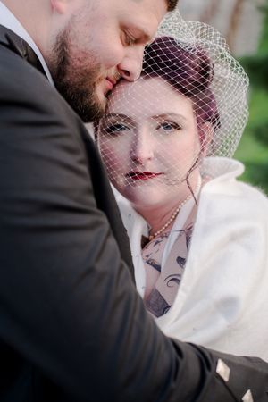 Groom tenderly kisses brides forehead as she gazes forward wearing birdcage veil and pearls, In Brussels Ontario.
