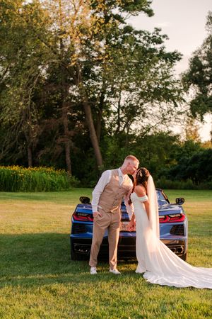 Bride and groom share a romantic kiss leaning against a blue sports car on a lush green lawn at golden hour.