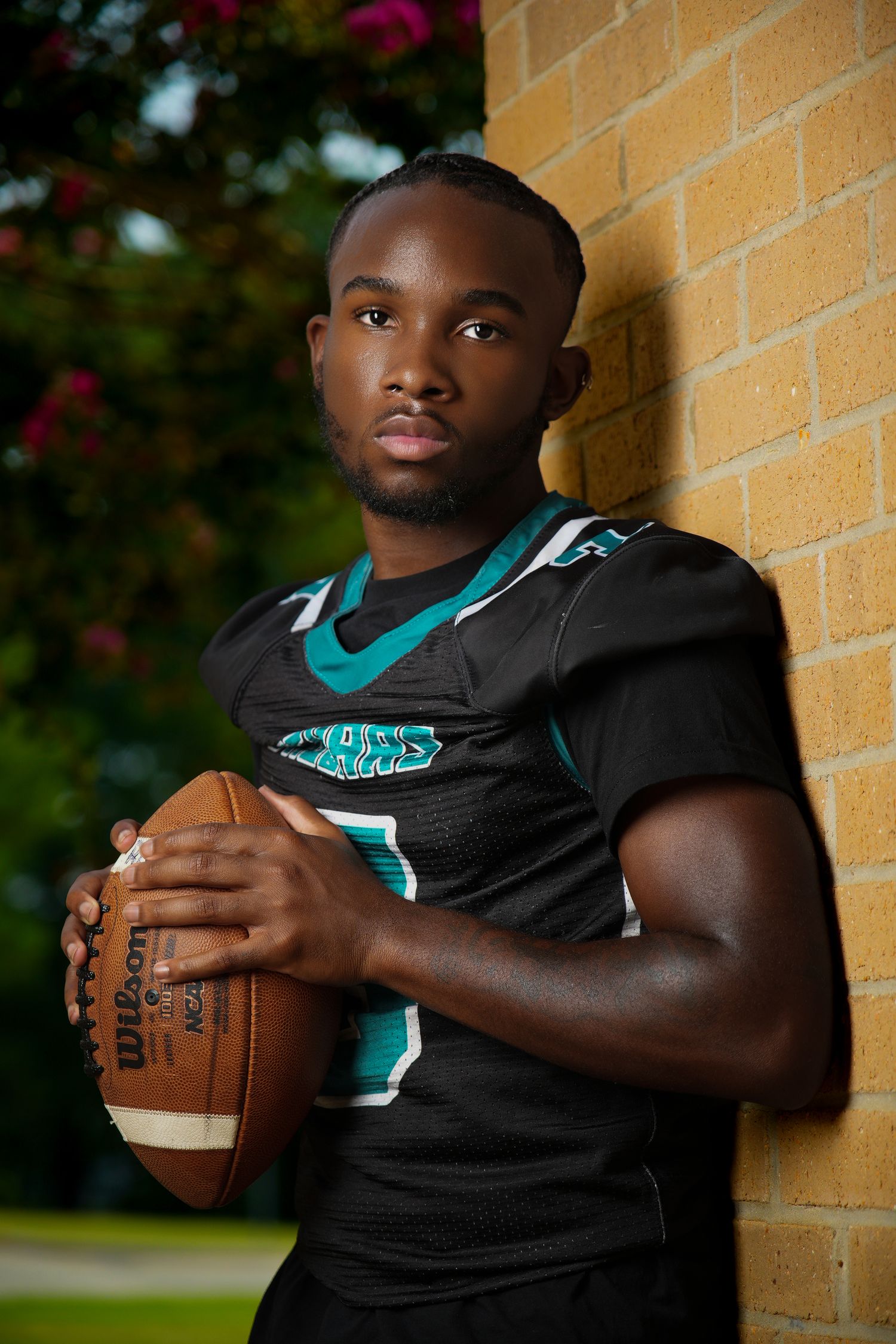 Football player in teal and black uniform poses against yellow brick wall holding game ball.
