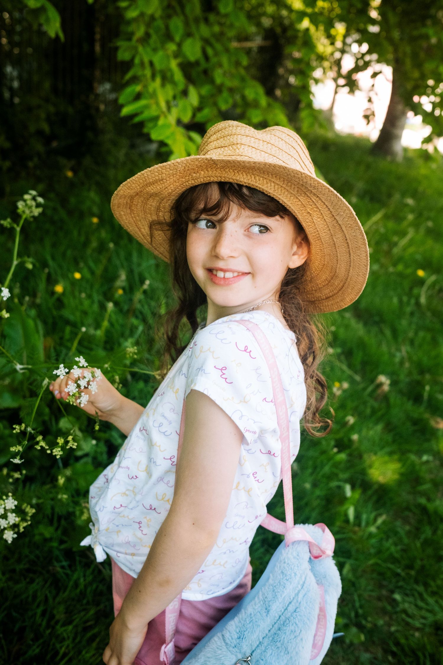 A person wearing a straw hat and white floral top poses outdoors among greenery on a sunny summer day.