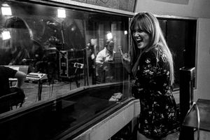 Singer Deidre Thornell performing in recording booth with band visible through glass window.