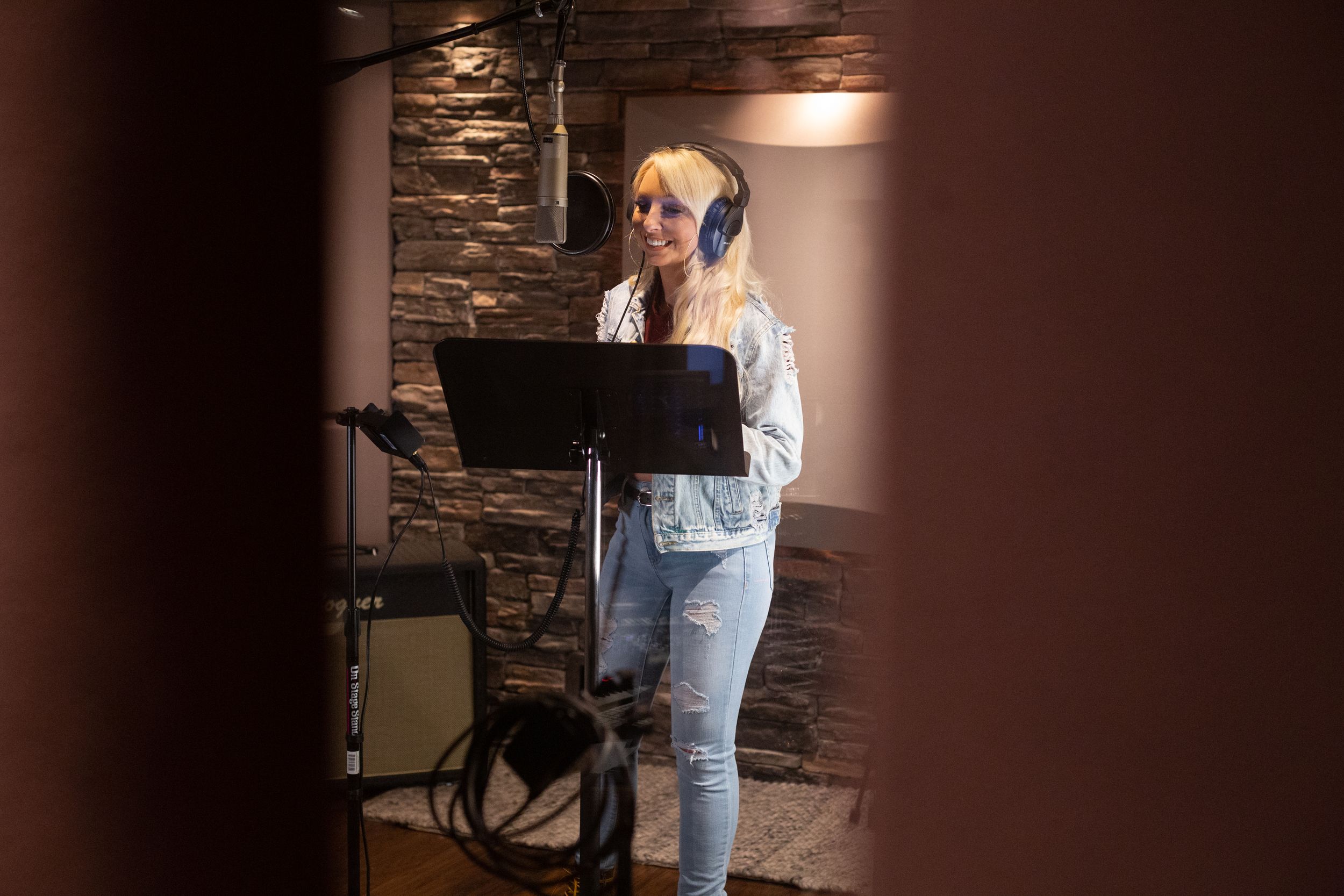 Singer Becca Bowen recording vocals in studio booth with music stand and stone wall background.