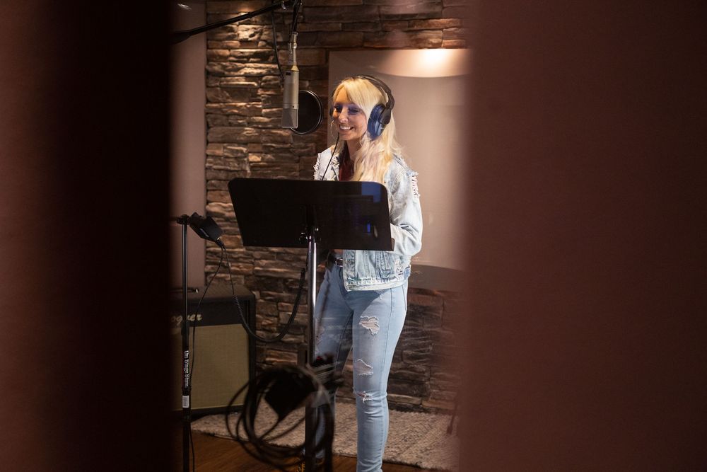 Singer Becca Bowen recording vocals in studio booth with music stand and stone wall background.