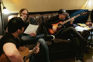 Musicians collaborating with acoustic guitars on leather couch during recording session.