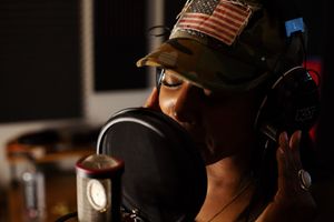 Recording artist Brei Carter in hat with American flag sings into professional microphone in dark studio setting.