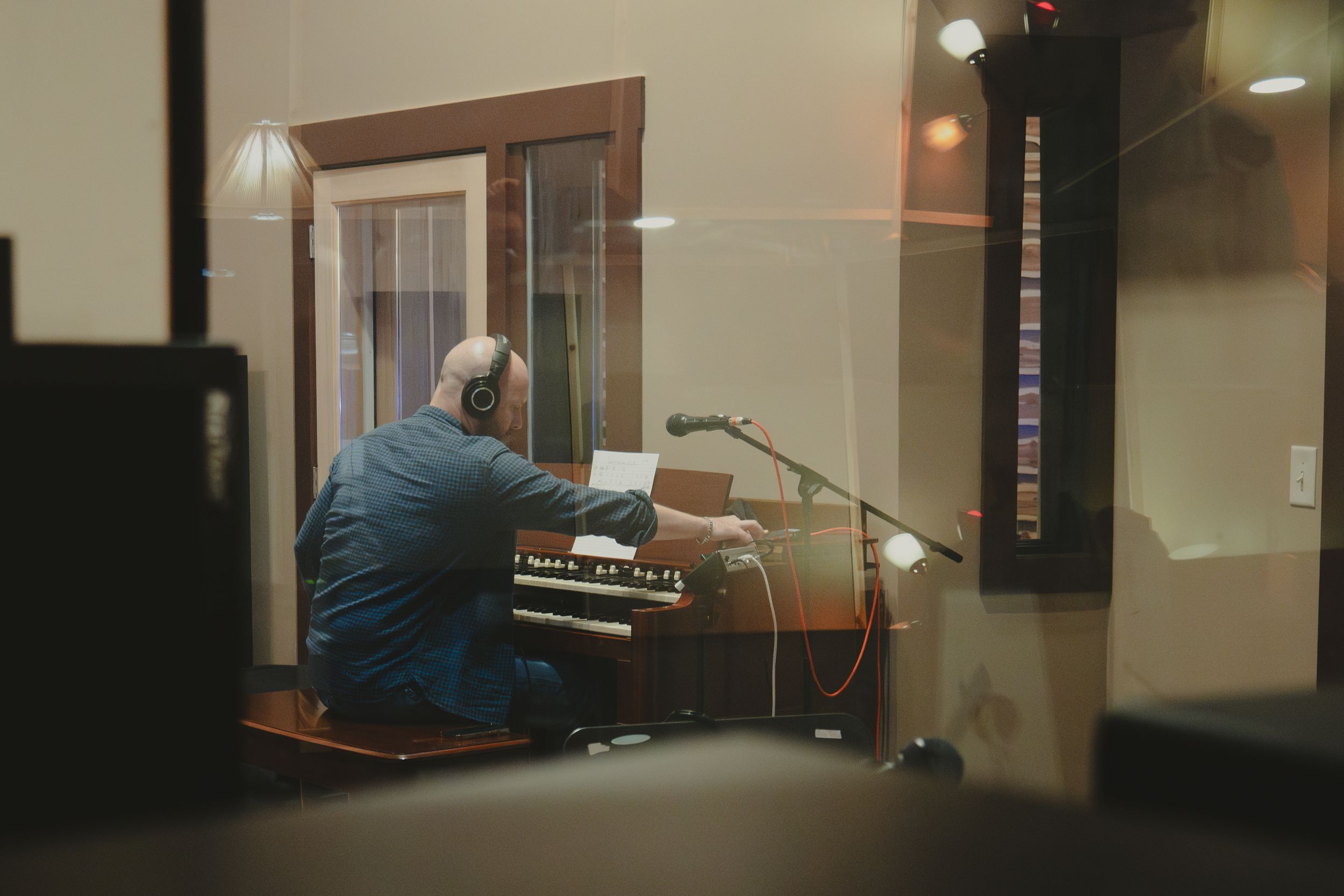 Person playing piano in isolated recording booth viewed through glass window.