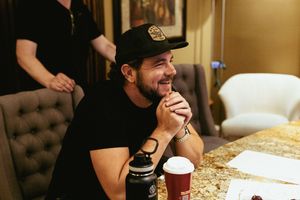 Music artist Matt Jordan wearing a baseball cap smiles while sitting at a table with a drink in a recording studio.