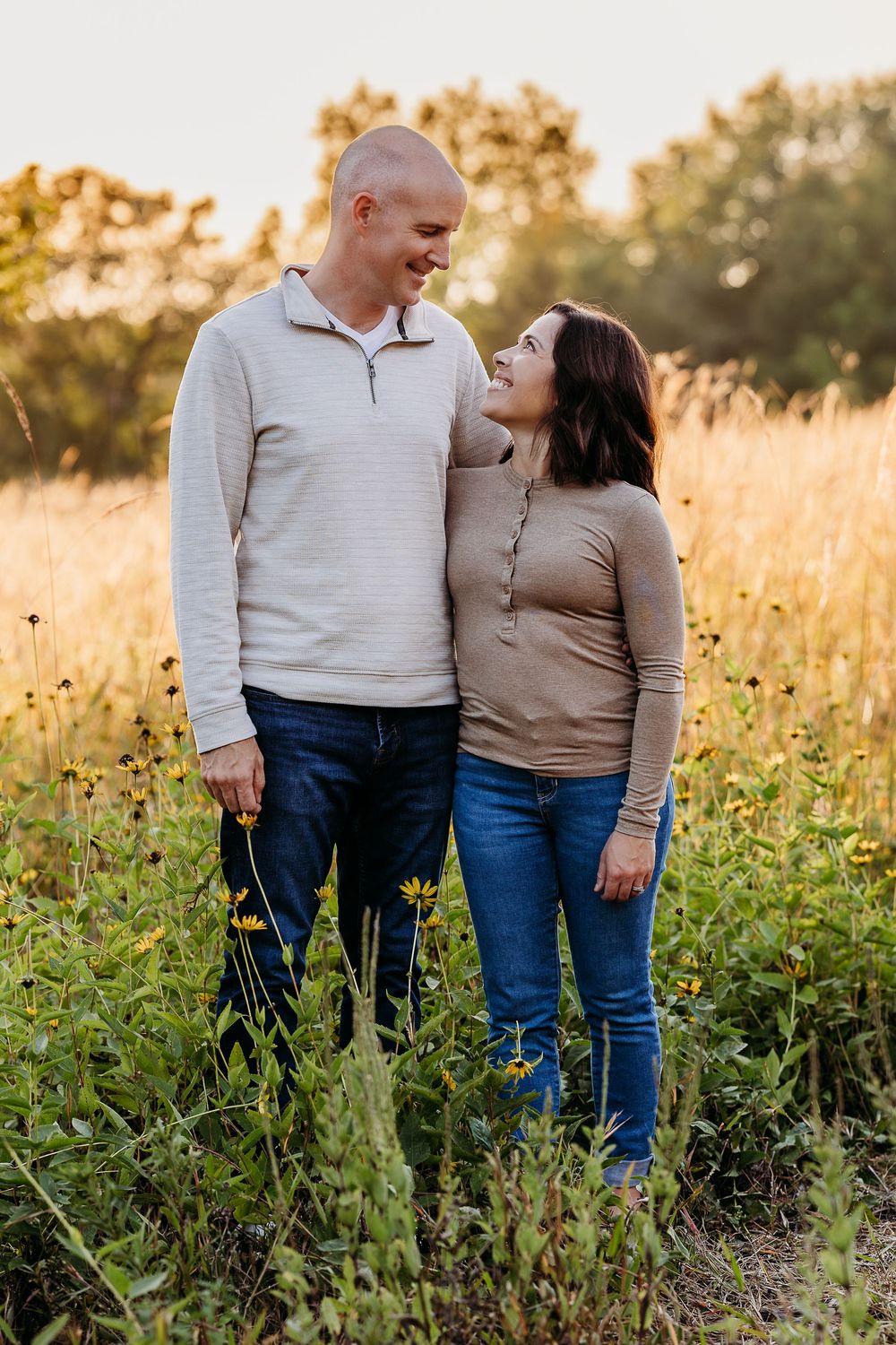 A couple stands together in a golden wildflower field during sunset wearing casual sweaters and jeans.