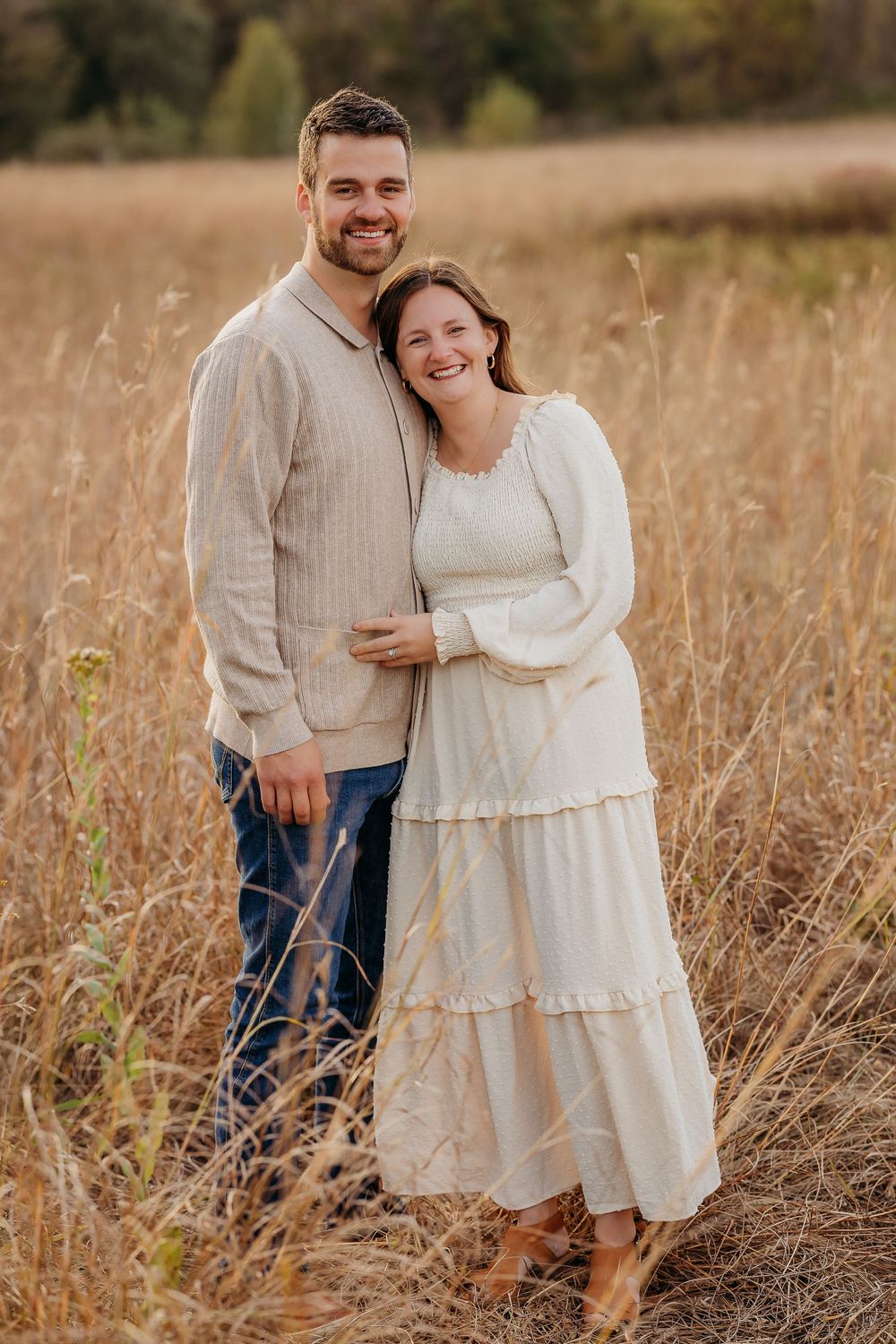A couple poses together in a golden field during sunset wearing coordinated cream and white outfits.