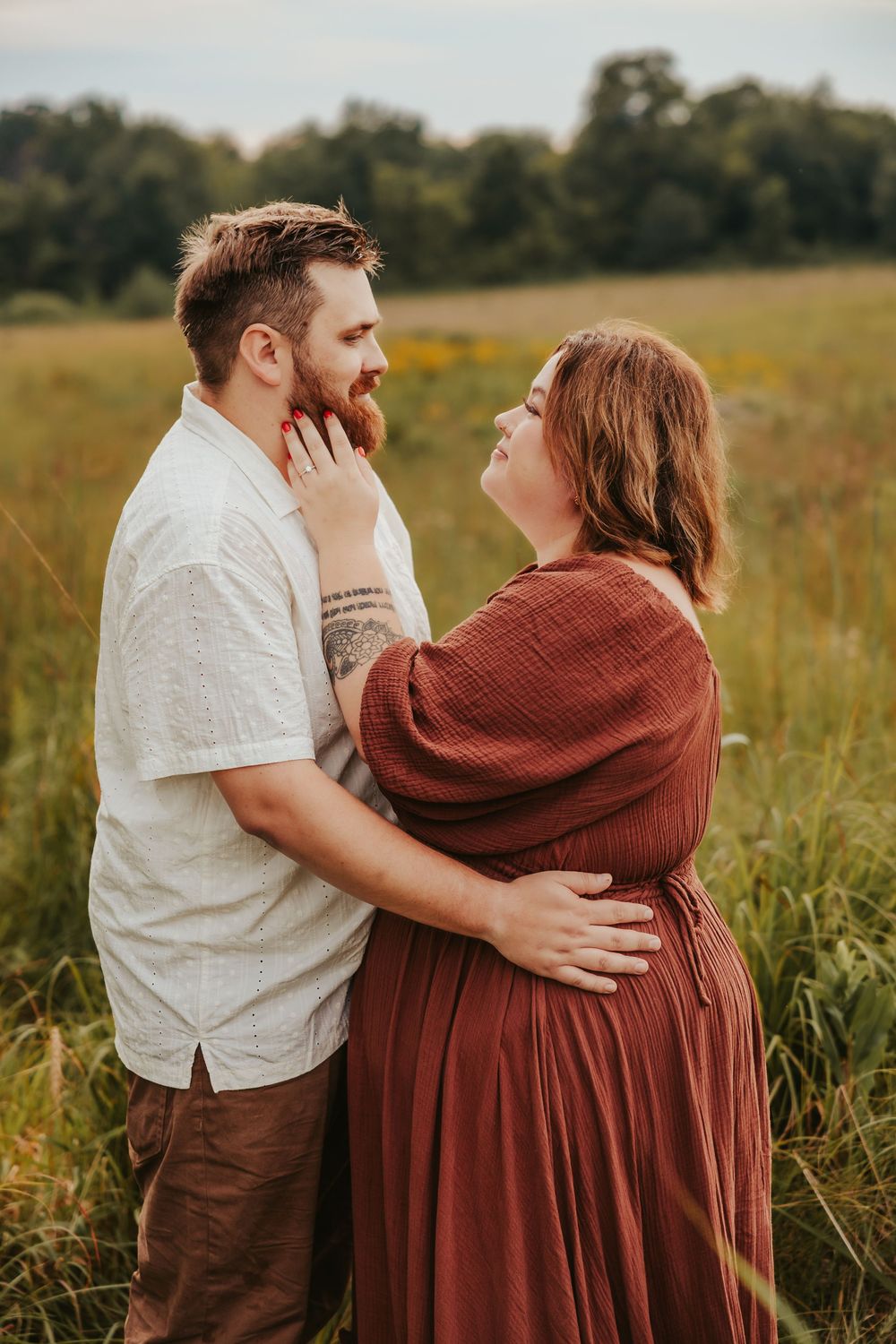 A tender moment between a couple standing in a golden field during sunset, she wearing a rust-colored dress.