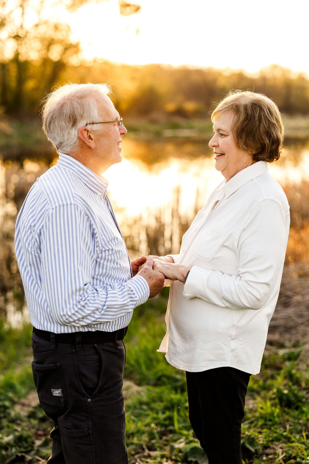 Elderly couple shares tender moment holding hands at sunset by a lake with golden light glowing behind them.