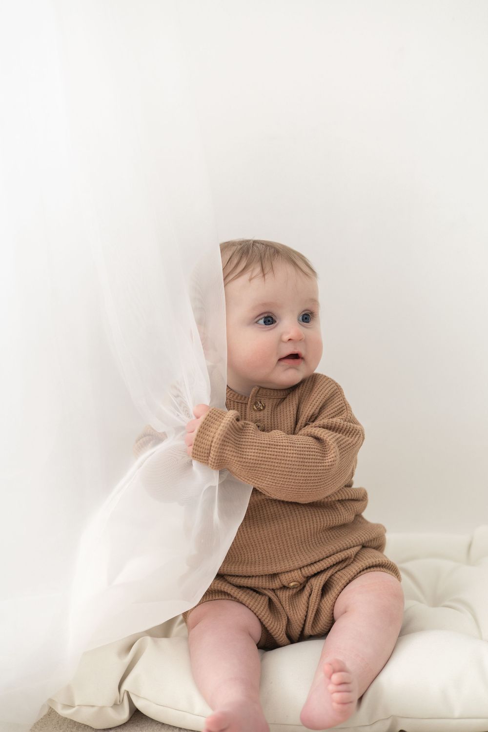 Cute baby in brown ribbed romper sitting on white cushion, holding sheer fabric curtain baby / milestone birthday photoshoot Nottingham