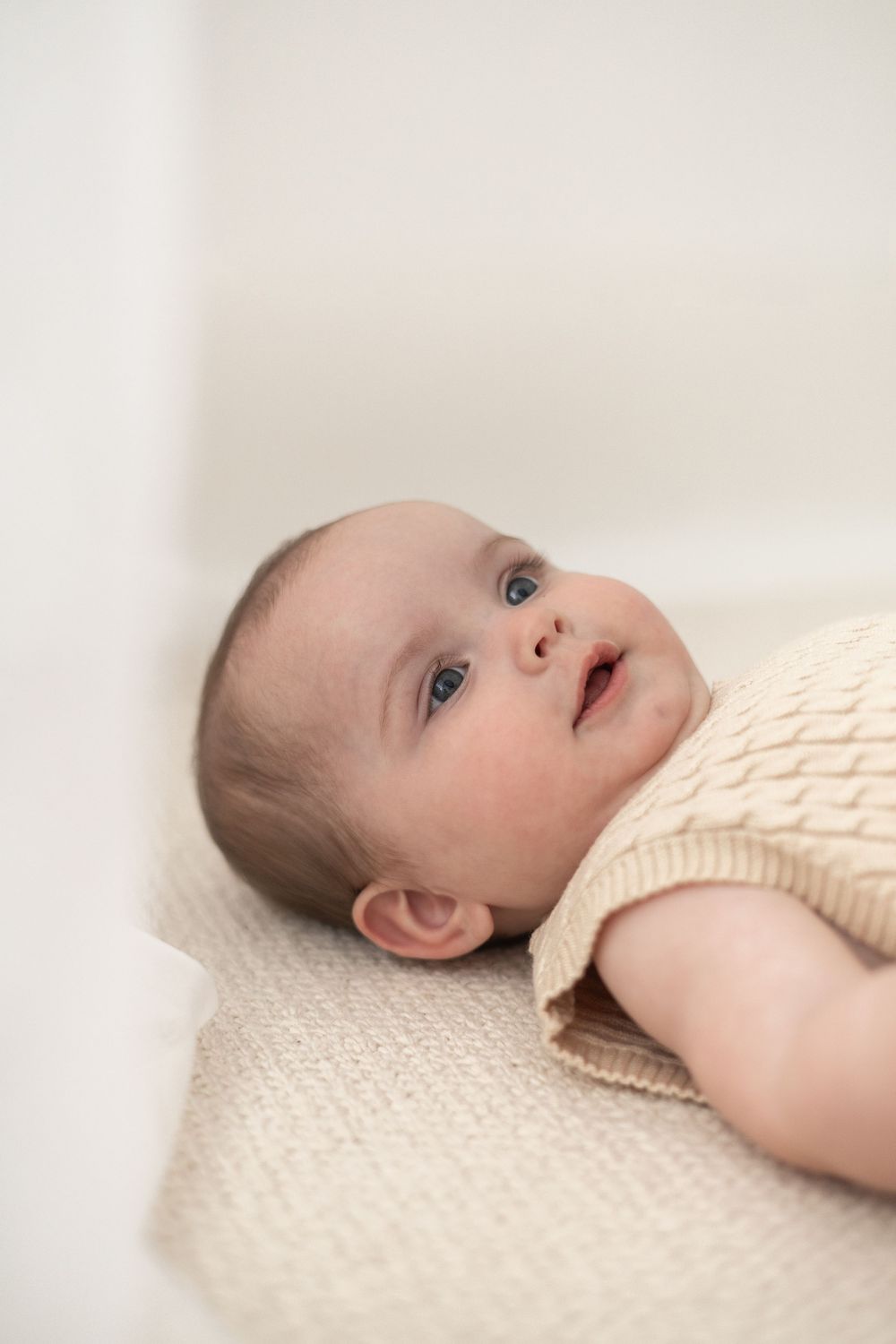 Baby lying on a cream knit blanket, wearing a beige sweater, looking up with bright eyes and a gentle smile milestone photoshoot Nottingham