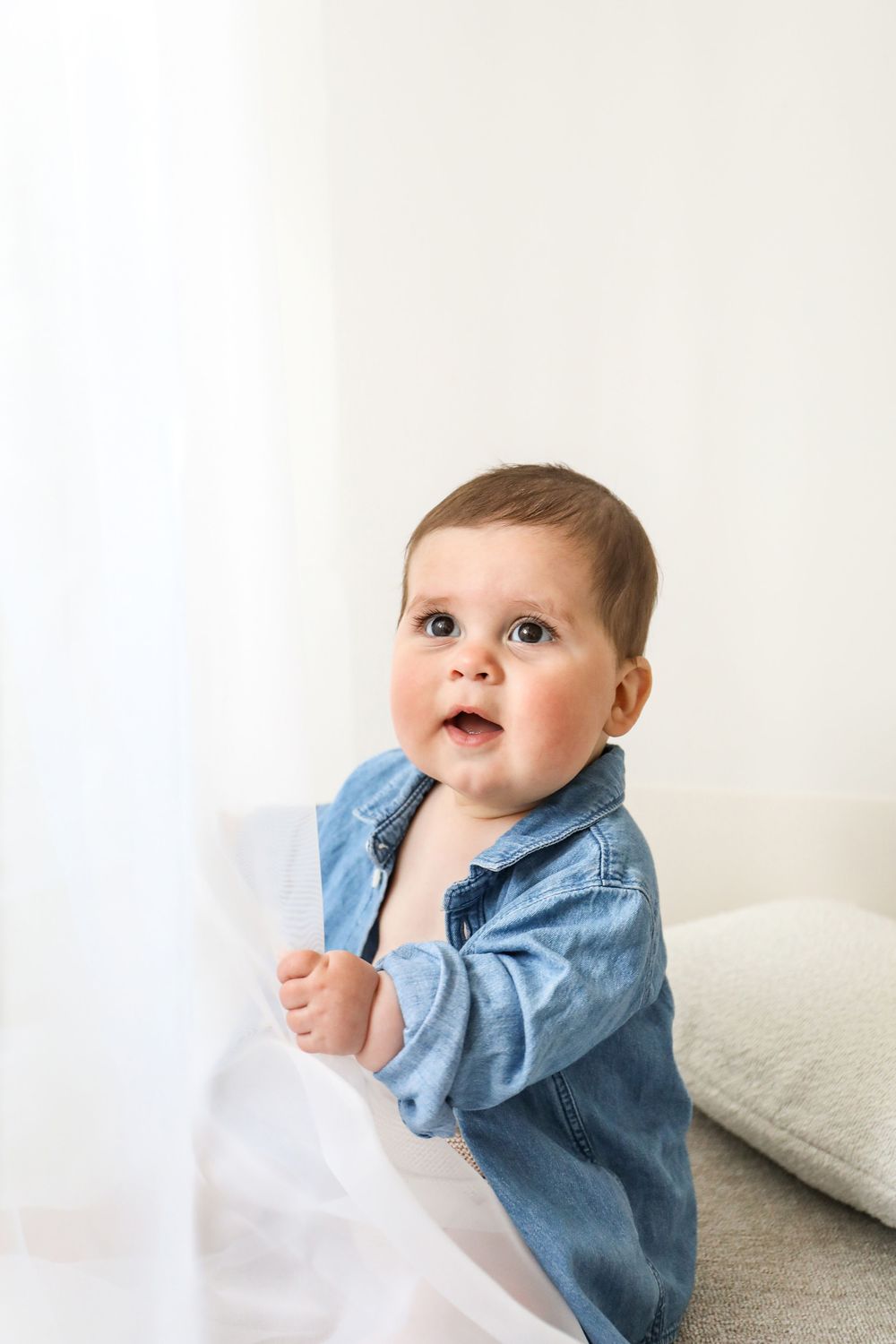 Infant in denim button-up shirt looking upward while sitting against white backdrop with pillows.