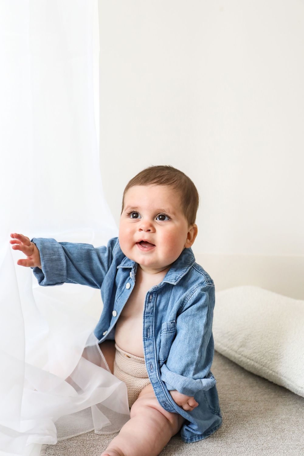 Baby wearing blue denim jacket reaches toward white curtain while sitting on couch.