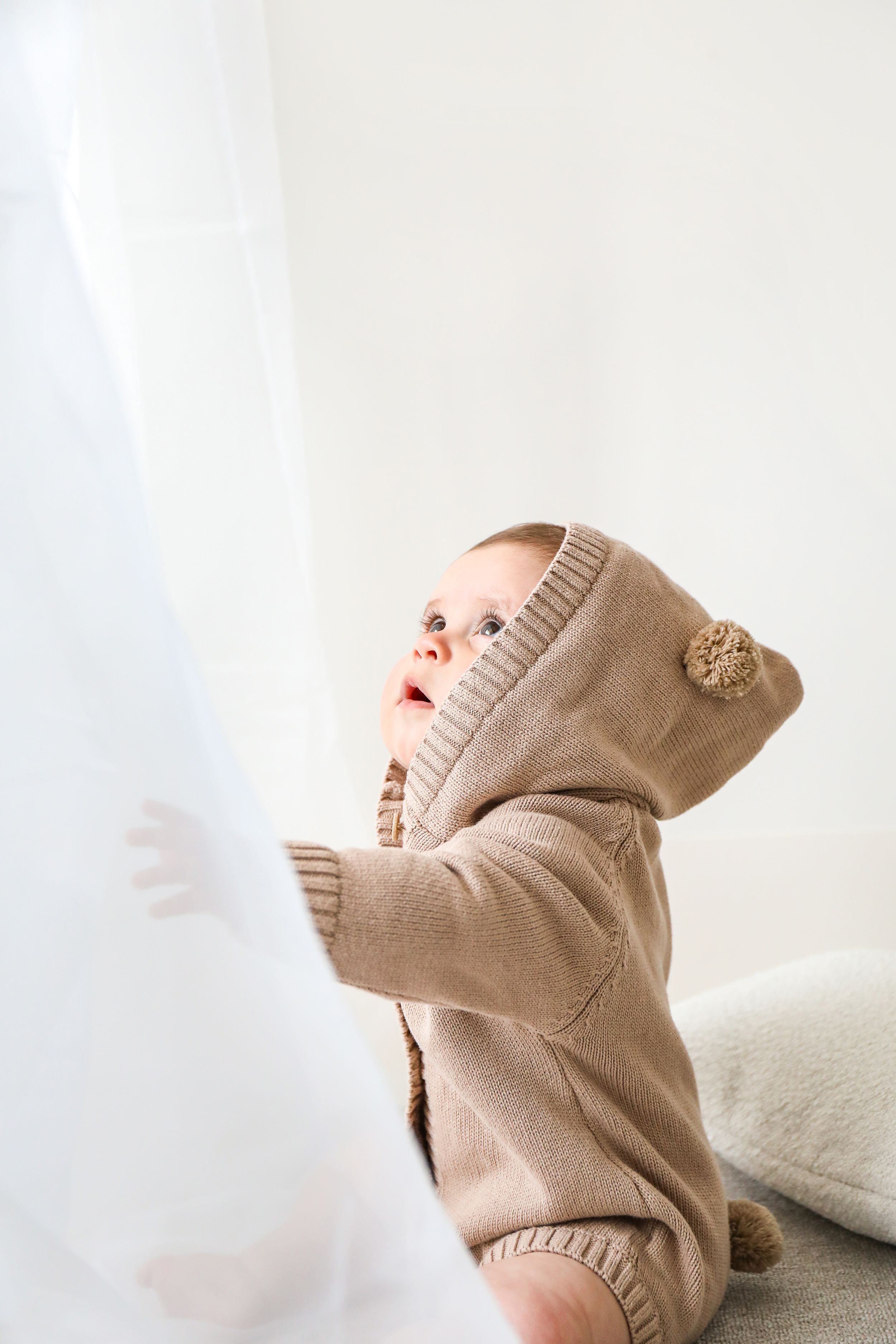 Infant wearing a cozy beige knit hooded sweater reaching up while sitting on white fabric.