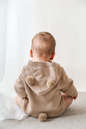 Baby wearing cozy beige knit jumper with pom pom details sits with their back to camera on light grey surface.