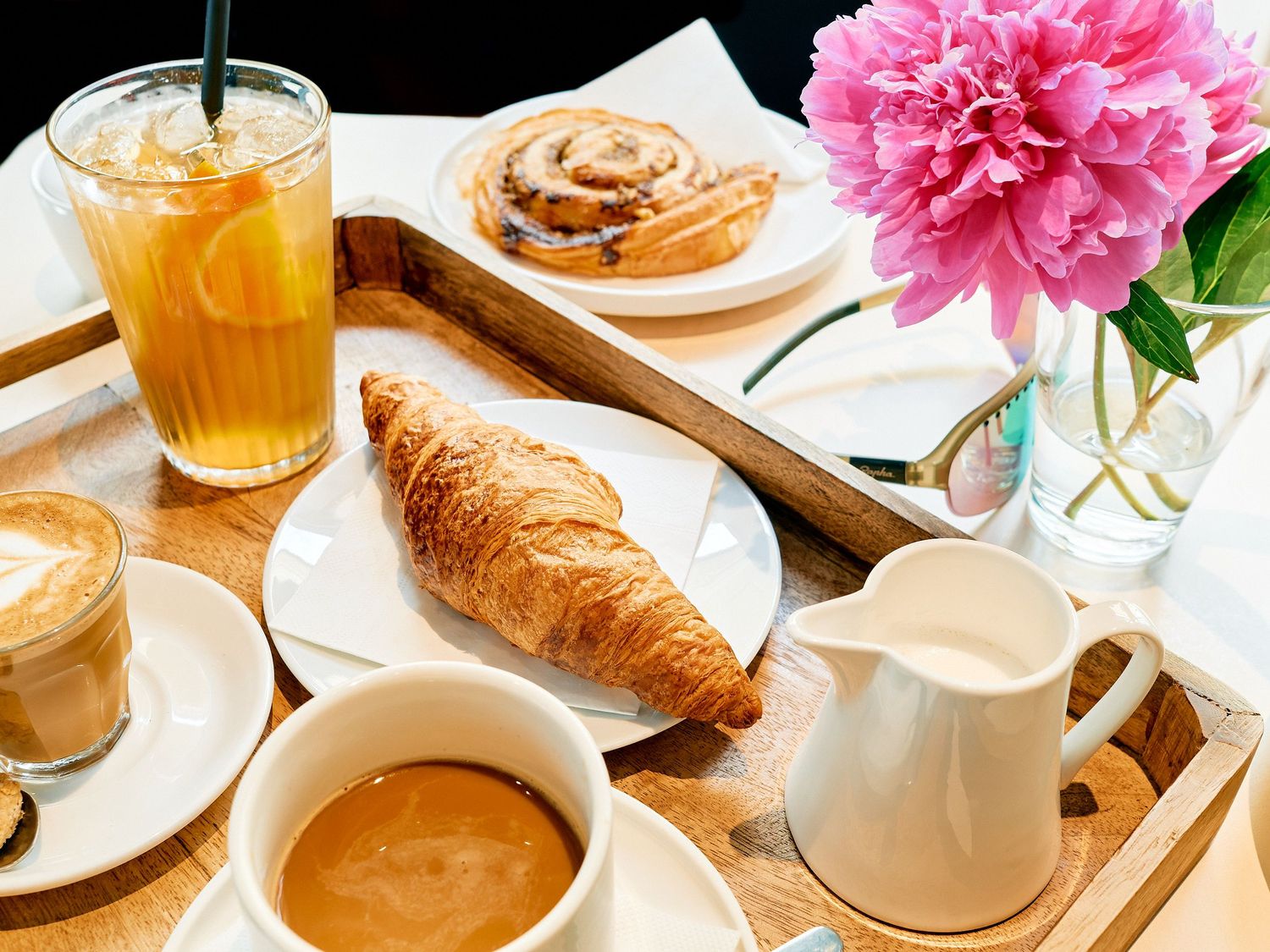 A breakfast spread with croissant, cinnamon roll, coffee, orange juice and pink peony flower in a vase on wooden tray.