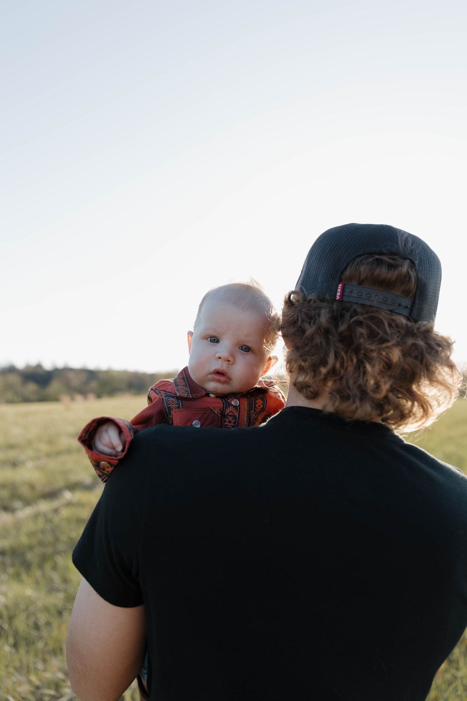 A parent in a black shirt and baseball cap holds a baby wearing plaid while standing in a sunlit meadow.