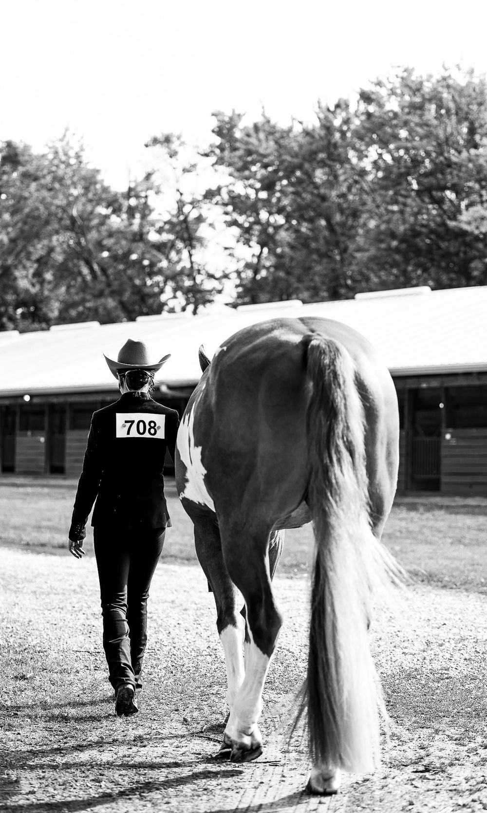 A cowboy in competition attire walks alongside a horse wearing number 708 near barn stables.