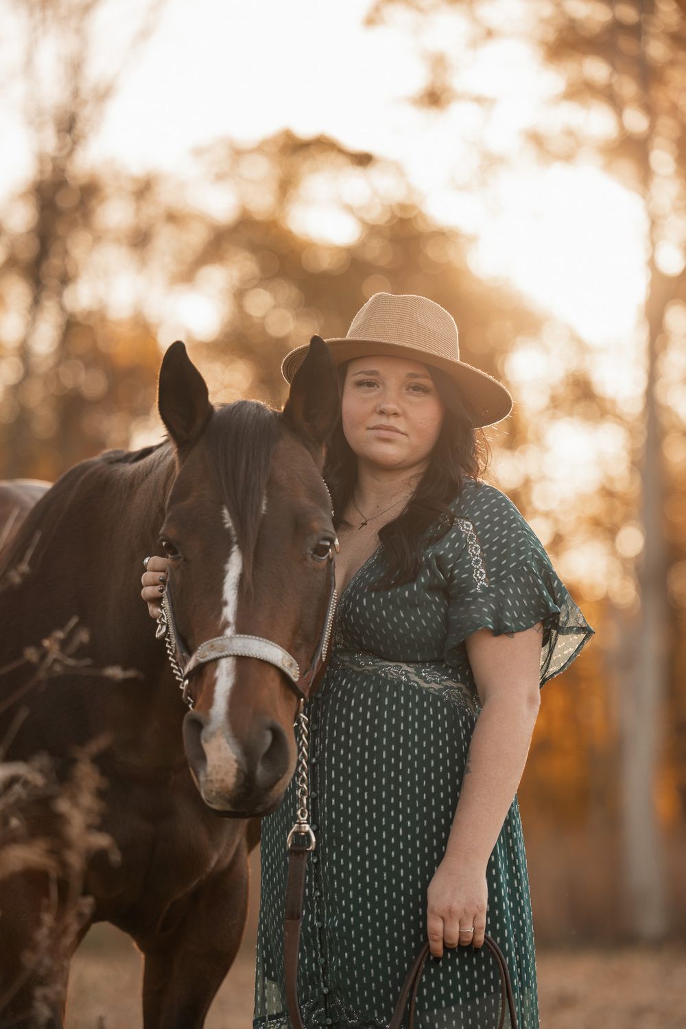 A woman in a patterned dress and hat stands with her dark horse at golden hour.